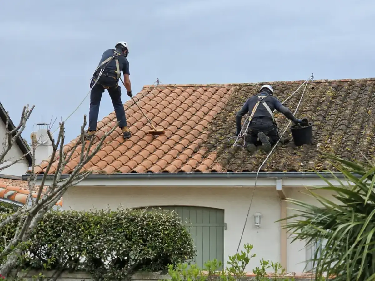 Toiture en tuile canal à moitié démoussée, deux couvreurs encordés au travail