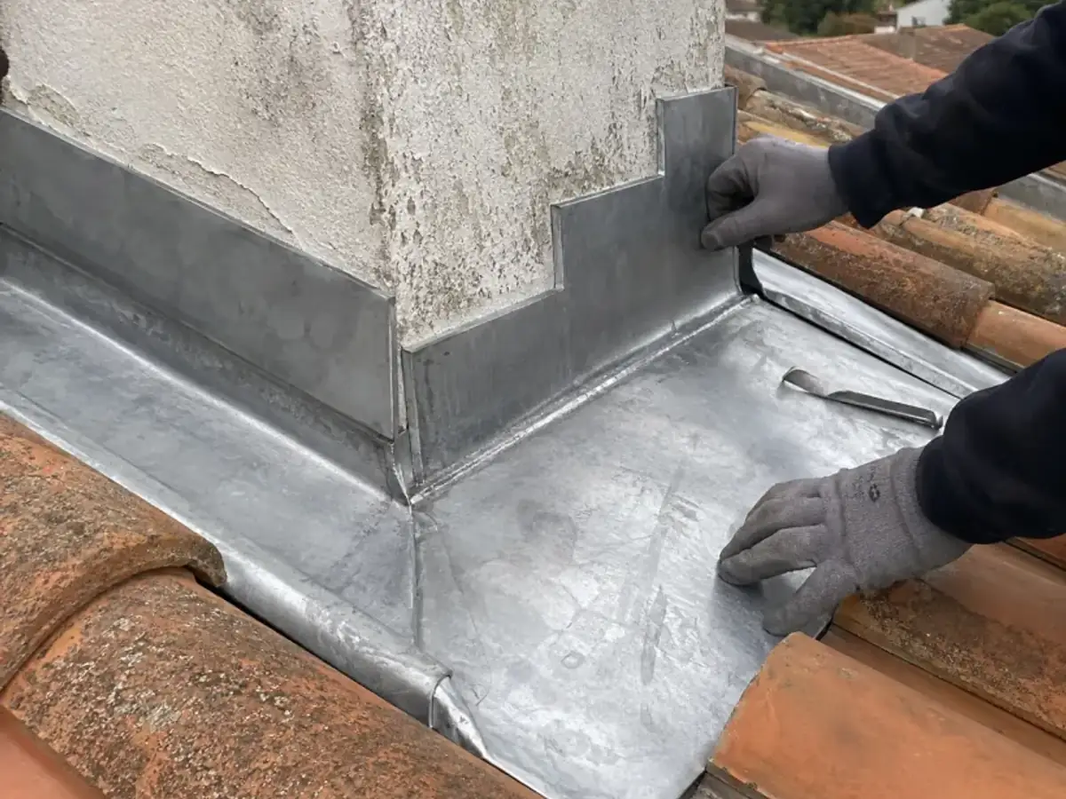 Mains gantées ajustant un abergement en zinc au pied d'une souche de cheminée, tuiles canal autour